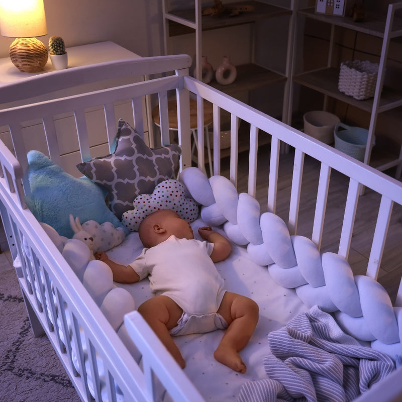 A baby doll lying on a white crib mattress with a white braided crib bumper, patterned pillows, and a stuffed bunny in a softly lit nursery.