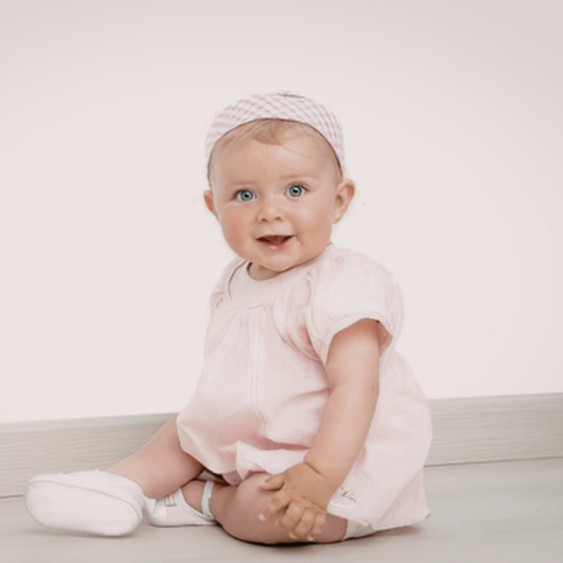 A baby sitting on a light floor wearing a pale pink short-sleeve romper, a matching pink and white checkered headband, and white booties against a plain off-white background.