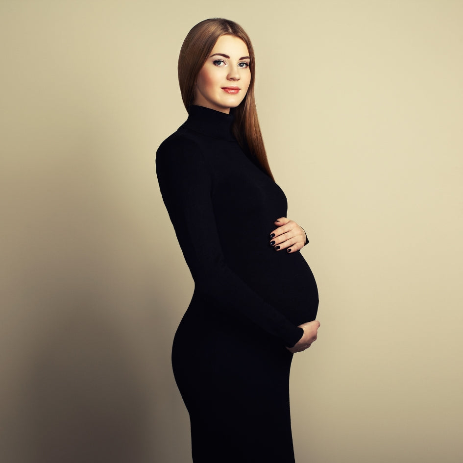 A pregnant woman with long light brown hair stands in profile wearing a fitted black long-sleeve turtleneck dress against a plain warm beige studio background.