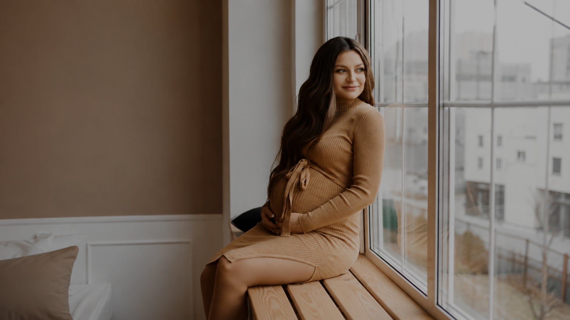 A pregnant woman with long dark hair sits on a wooden window ledge wearing a fitted tan maternity dress, with a softly lit room and large windows in the background.
