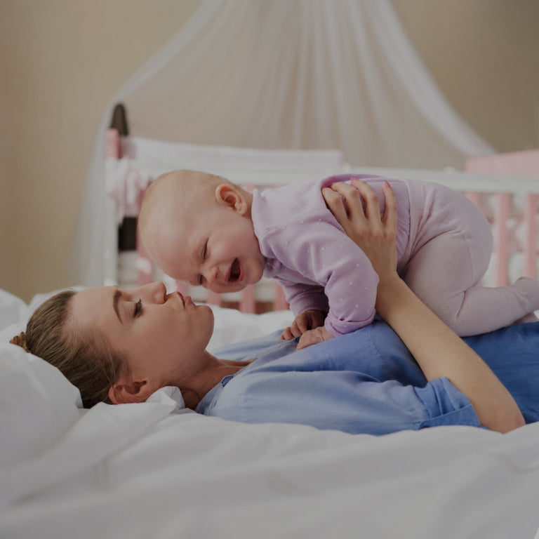 A woman lying on a bed with white bedding holds and kisses a smiling baby wearing a light purple onesie in a softly lit bedroom.