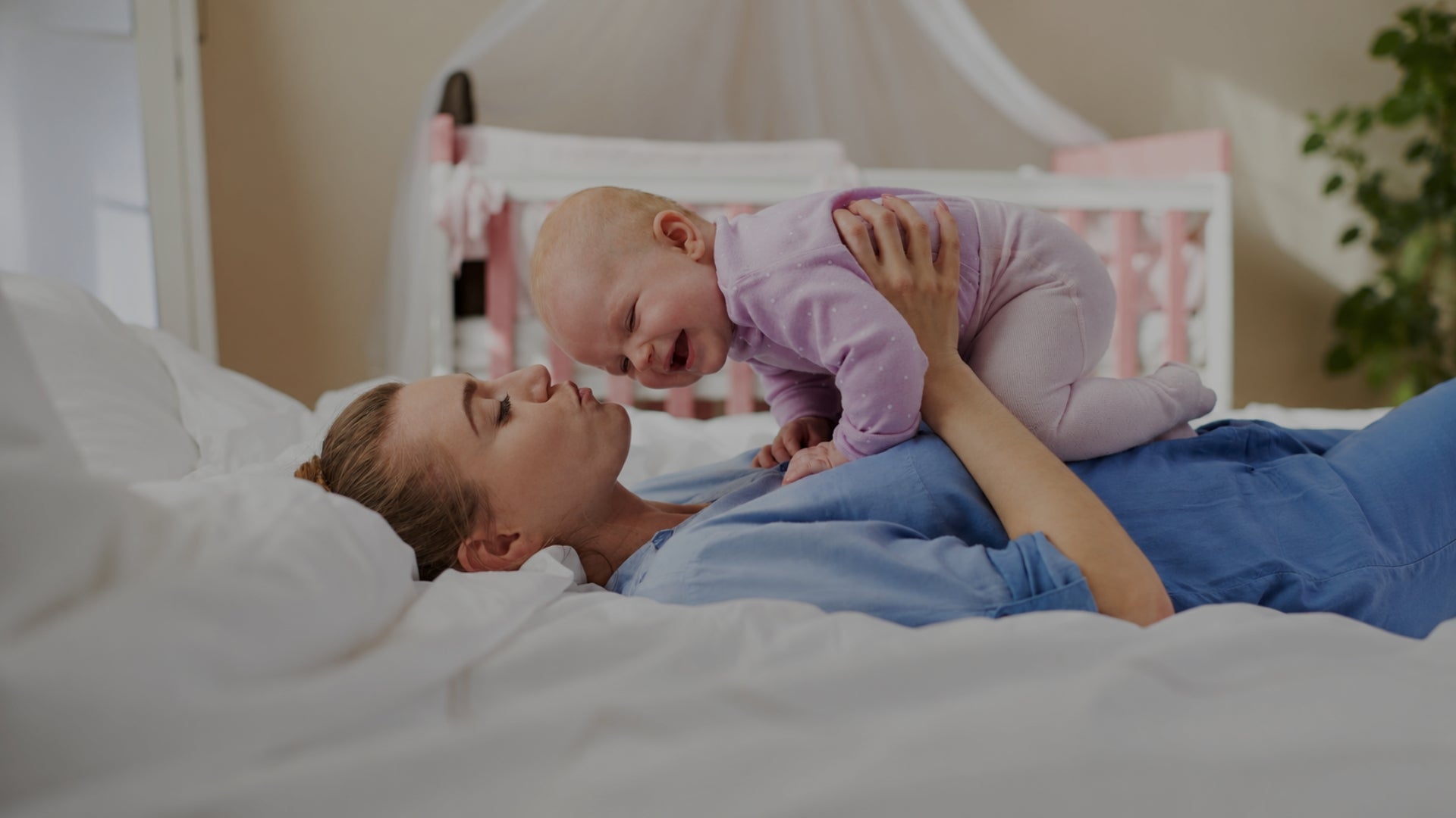 A woman lying on a bed with white bedding holds and kisses a smiling baby wearing a light purple onesie in a softly lit bedroom.